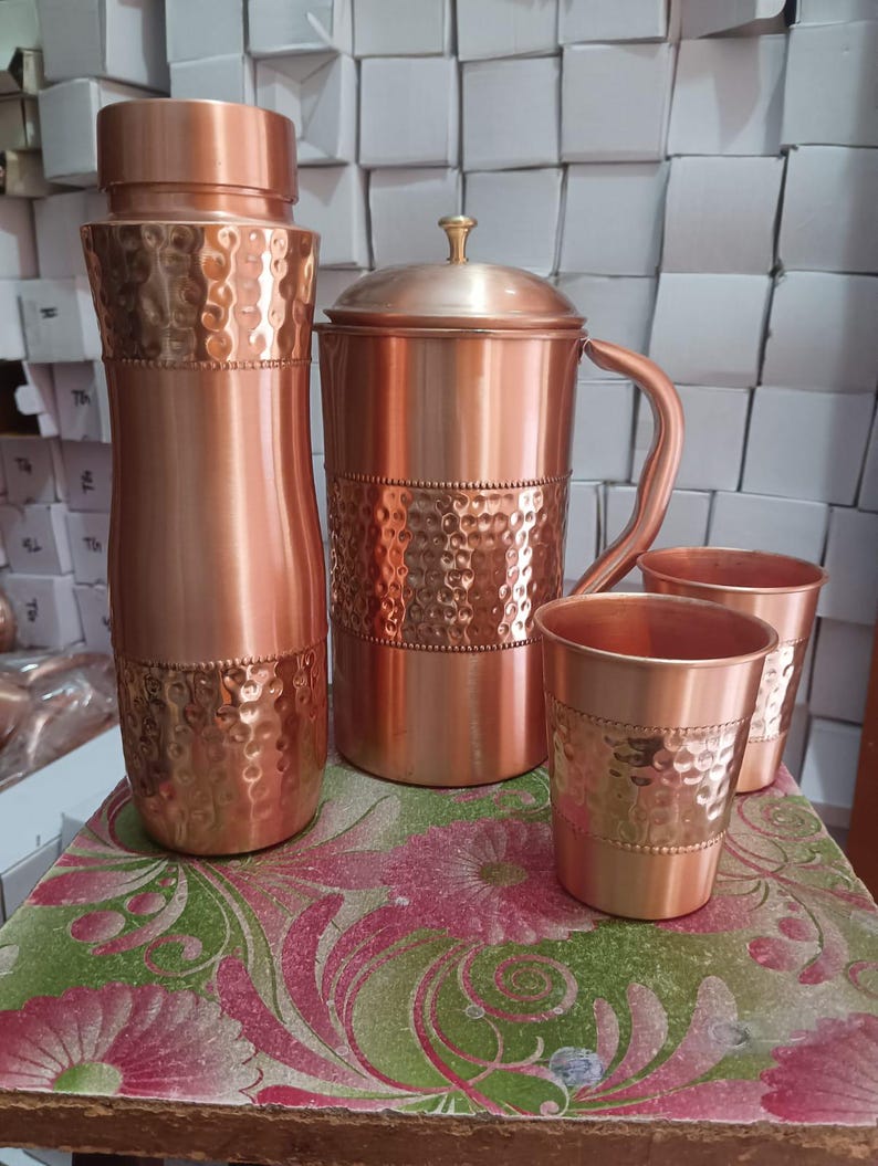 May include: A set of copper drinkware consisting of a tall water bottle, a pitcher with a lid and handle, and two tumblers. Each item features a polished finish with a central band of hammered texture and a decorative beaded border detail.