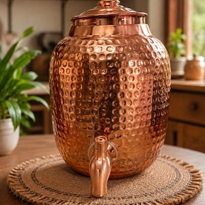 May include: A 4 litre copper dispenser with a hammered texture, a matching lid with a small knob handle, and a front-facing spigot. The text 4 litre dispenser available New look is displayed at the top of the image in yellow and white.