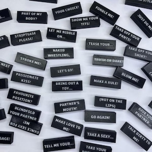 Könnte beinhalten: Eine Ansammlung schwarzer, rechteckiger Blöcke mit weißer Schrift, die auf einer weißen Oberfläche verstreut sind. Die Blöcke zeigen verschiedene anzügliche Sätze, darunter "Stroke Me!", "Let's 69!" und "Take Off Your Pants!". Artikel für erotische Spiele.