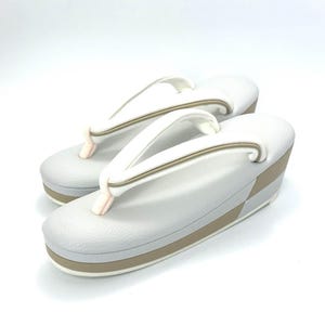May include: A pair of white zori sandals featuring a textured surface, a raised platform sole with a tan horizontal stripe, and white thong straps with a subtle tan trim. The sandals have a rounded toe design and a light pink connector piece.