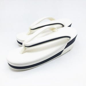 May include: A pair of white zori sandals featuring a thick platform sole with a dark navy blue accent stripe along the side. The thong-style straps are also white with a thin navy blue trim, designed for a secure fit on the footbed of the sandal.