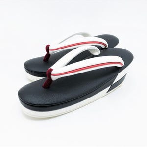 May include: A pair of zori sandals featuring a black textured footbed and a thick white platform sole. The thong straps are white with a thin red accent line and a dark maroon velvet-like toe post, creating a structured and elevated footwear design.