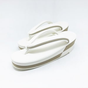 May include: A pair of white zori sandals featuring a thick, elevated sole with a light beige side panel. The thong-style straps are white with a subtle beige trim, designed with a rounded toe and a sturdy, platform-style base for support.