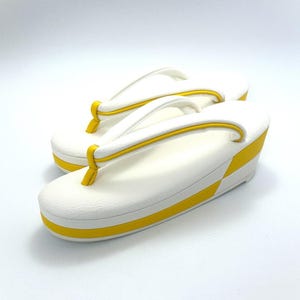May include: A pair of white zori sandals featuring a thick platform sole with a bright yellow horizontal stripe along the side. The thong-style straps are white with yellow piping, and the toe post is yellow, creating a clean, two-tone aesthetic.