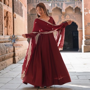 Op de afbeelding: Een bordeauxrode jurk met bijpassende sjaal. De jurk heeft lange mouwen en een zwierige rok. De sjaal is versierd met ingewikkelde gouden borduursels langs de randen. Het logo "NILESH FABRICS" is ook zichtbaar.