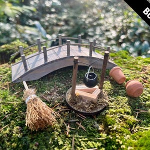 Puede incluir: Conjunto de jardín de hadas en miniatura con un puente de madera, una escoba, un caldero, dos macetas de terracota y un pozo. La escena está ambientada sobre un lecho de musgo verde, creando un paisaje en miniatura.