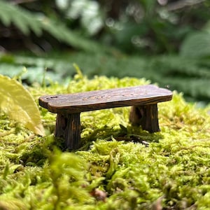Op de afbeelding: Een miniatuur houten bankje met een getextureerd oppervlak, rustend op een bed van groen mos. De bank heeft een bruine afwerking en is ontworpen met twee stevige poten. Een gele blad is zichtbaar op de voorgrond, met een vage groene achtergrond.