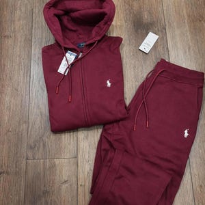 Puede incluir: Un conjunto de chándal burdeos, que incluye una sudadera con cremallera y pantalones de chándal a juego, se muestra sobre una superficie de madera. La sudadera con capucha tiene una capucha con cordón y un pequeño logotipo blanco. Los pantalones tienen cintura con cordón y un logotipo a juego.