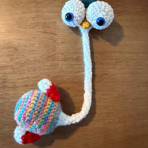 May include: A quirky, handmade crochet bookmark in the form of a cartoon character. It has two large, white, crocheted eyes with blue pupils, a white stem, and a colourful, striped body with red accents. The bookmark is designed to be placed in a book.