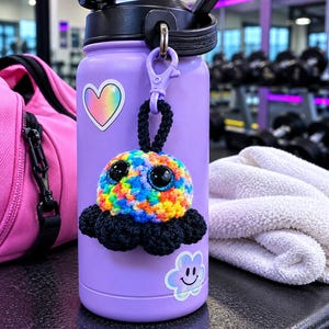 May include: A light purple water bottle featuring a rainbow-colored crochet octopus keychain with large black eyes and black tentacles. The bottle is decorated with a heart sticker and a smiling flower sticker, with a black lid and handle.