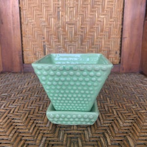Peut inclure: Un pot de plante carré en céramique vert clair avec une soucoupe assortie. Le pot présente une surface texturée de petits points en relief. Le design est une version moderne d'une forme classique, adaptée aux petites plantes.