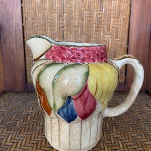 May include: A decorative ceramic jug with a colourful floral design. The jug features a cream-coloured base with vertical lines and a handle. The upper portion is adorned with red, yellow, blue, and orange leaf-like shapes.