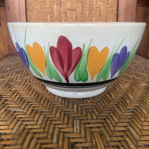 May include: Cream-colored ceramic bowl with a floral design. The bowl features colorful painted flowers in red, orange, and purple, with green leaves. A black stripe encircles the bowl near the top, and it sits on a white base.