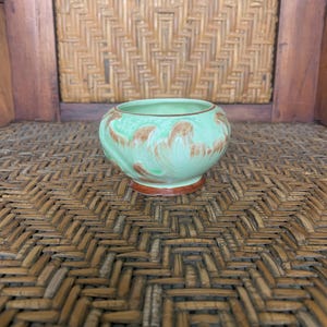 May include: A small, round, ceramic bowl with a light green glaze and brown brushstroke design. The bowl has a brown base and rim. The background is a woven pattern.