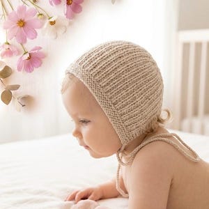 Puede incluir: Un gorro de bebé tejido de color beige con correas. El gorro está hecho de una tela de punto acanalado. Flores rosas y blancas y vegetación en el fondo. El bebé está acostado sobre una superficie blanca.
