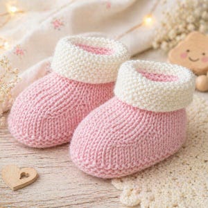 May include: A pair of pink and white knitted baby booties. The booties have a ribbed texture and a folded-over cuff. A wooden heart and a baby rattle are in the background.