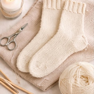May include: A pair of cream-colored, hand-knitted socks rest on a beige fabric, alongside a ball of yarn, knitting needles, and a lit candle in a glass jar. Silver scissors are also present, suggesting a crafting theme.