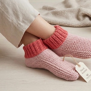 May include: A pair of hand-knitted ankle socks featuring a light pink textured knit body and a contrasting ribbed coral-colored cuff. A small beige paper tag attached with twine to the front sock displays the text Hand-Knitted in black ink.