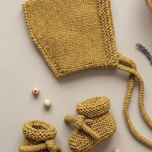 May include: Mustard yellow knitted baby bonnet and booties set. The bonnet has tie strings, and the booties have a folded cuff. The set is made of a chunky knit yarn. Wooden beads are scattered around the set.