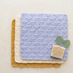 May include: Three knitted washcloths in shades of yellow, cream, and light blue are stacked. A bar of soap and two green eucalyptus leaves rest on the blue cloth. The washcloths have a textured, geometric pattern.