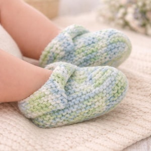 Pode incluir: Um par de botinhas de bebé feitas à mão, em tons pastel de azul e verde. As botinhas têm um aspeto texturado e estão colocadas sobre uma manta de malha creme. As cores suaves e a textura de malha sugerem um design acolhedor.