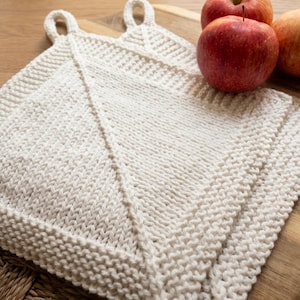 Può includere: Due presine quadrate in maglia bianco sporco con manici ad anello sono esposte su una superficie di legno, accanto a tre mele rosse. Le presine hanno un aspetto strutturato e un elemento di design diagonale.