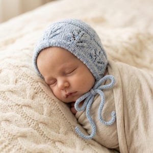 Könnte beinhalten: Ein schlafendes Baby mit einer hellblauen Strickhaube mit einem zarten Blättermuster und einer passenden Schleife. Das Baby ist in eine weiche, cremefarbene Decke gehüllt und liegt auf einer strukturierten Zopfstrickdecke.
