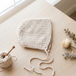 May include: A beige, hand-knitted baby bonnet with tie strings, lying on a light wooden surface. A ball of yarn and a knitting needle are next to the bonnet. A dried rose and sprig of greenery are also on the surface.
