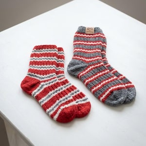 May include: A pair of knitted socks featuring horizontal stripes in red, grey, and white. One sock is primarily red with grey and white stripes, while the other is primarily grey with red and white stripes. A small tan tag with a ram logo and the text ANTWYLL is attached to the cuff of the grey sock.