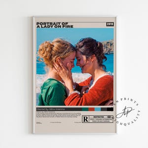May include: A framed poster featuring two women embracing, with the title "Portrait of a Lady on Fire" at the top. One woman wears a green dress, the other a red top. The background shows a blue sea and sky.