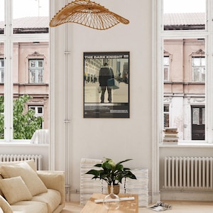 May include: A framed movie poster titled The Dark Knight 2008 featuring a central graphic of a person standing in a city street, rendered in muted tones of beige, gray, and black, displayed on a white wall above a wooden table and a green plant.