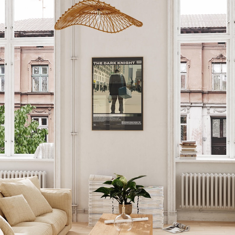 May include: A framed movie poster titled The Dark Knight 2008 featuring a central graphic of a person standing in a city street, rendered in muted tones of beige, gray, and black, displayed on a white wall above a wooden table and a green plant.