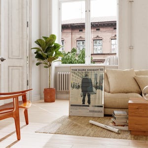 May include: A framed movie poster for The Dark Knight featuring a central figure walking through a city street, with the film title in bold black text at the top and a color palette strip at the bottom, displayed in a bright living room.