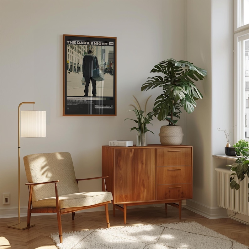 May include: A framed movie poster titled The Dark Knight featuring a figure in a dark coat walking through a city street, displayed on a wall above a wooden cabinet with three drawers and a door, next to a beige armchair and a tall floor lamp.