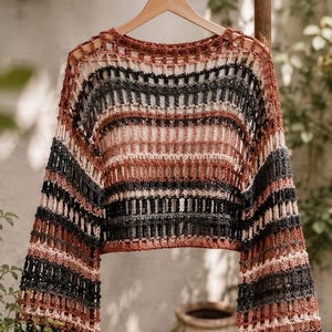 Könnte beinhalten: Ein gecroppter Pullover mit offenem Webmuster in Schwarz, Braun und Rosa. Das langärmlige Oberteil ist im Häkelstil gestrickt und hängt an einem Holzbügel. Der Pullover hat ein gestreiftes Muster und wird im Freien präsentiert.
