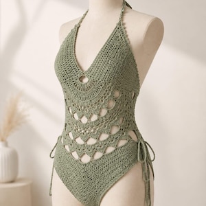 May include: A sage green crochet one-piece swimsuit featuring a halter neckline, a deep V-neck, and an intricate openwork pattern across the torso. The garment includes adjustable side ties at the hips and a high-cut leg design on a mannequin.