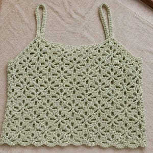 May include: A light green crochet top with a floral pattern. The top has thin straps and a scalloped hemline. The handmade garment is displayed on a neutral-colored surface.