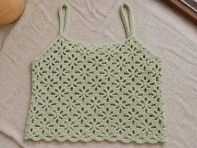 May include: A light green crochet top with a floral pattern. The top has thin straps and a scalloped hemline. The handmade garment is displayed on a neutral-colored surface.