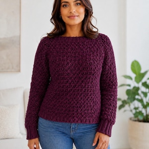 May include: A deep plum-colored long-sleeved sweater featuring a textured, repeating knit pattern, a round neckline, and ribbed cuffs at the wrists and hem, paired with classic blue denim jeans. The garment has a consistent, dense stitch design.