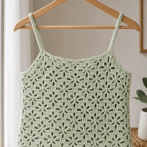 Pode incluir: Top de crochê verde claro com um padrão floral, pendurado em um cabide de madeira. O top apresenta uma borda com vieiras e alças finas. A peça de roupa feita à mão é exibida em um ambiente bem iluminado.