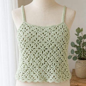 May include: A light green crochet top with a floral pattern. The top has thin straps and a scalloped hem. The garment is displayed on a mannequin.