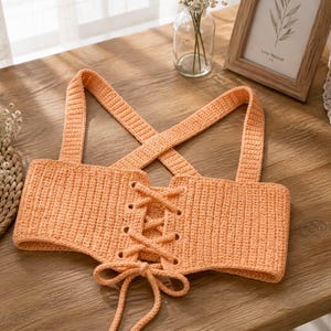 May include: Peach-colored crochet top with a corset-style front and crisscross straps. The top features a lace-up design in the center and is made of a textured, knitted material. The straps cross in the back.