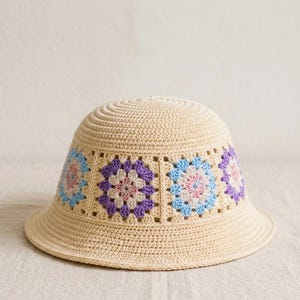 Può includere: Un cappello a secchiello all'uncinetto color crema con un motivo floreale. Il cappello presenta un'ampia tesa e una corona arrotondata. Il motivo floreale include quadrati con fiori blu, rosa e viola. Il cappello è realizzato in filato morbido e strutturato.