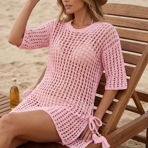 May include: A light pink crochet cover-up dress with short sleeves and side ties. The dress is worn with a straw hat. The open weave design is visible. The model is sitting on a wooden beach chair.