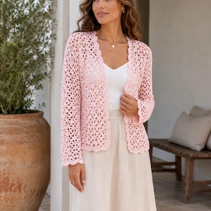 May include: A light pink open-front cardigan featuring a delicate crochet pattern with long sleeves and a scalloped edge trim. The garment is worn over a simple white camisole and a plain beige skirt, creating a layered look with a textured finish.