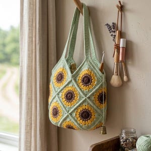 May include: A light green crochet tote bag with a sunflower design in yellow and brown. The bag is suspended from a wooden hook. A small brass bell hangs from the bag. The bag is handmade.