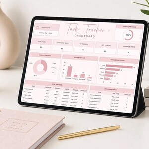 May include: A tablet displaying a task tracker dashboard in pink and white. A notebook with the text "Focus Plan Achieve" and a gold pen are in the foreground. A beige vase and a cup are in the background.