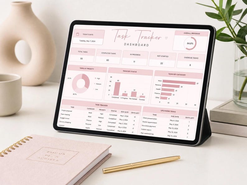 May include: A tablet displaying a task tracker dashboard in pink and white. A notebook with the text "Focus Plan Achieve" and a gold pen are in the foreground. A beige vase and a cup are in the background.