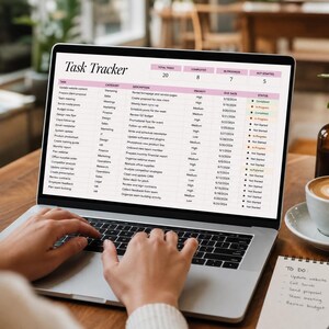 May include: A laptop screen showing a "Task Tracker" spreadsheet with tasks, categories, and deadlines. A coffee cup with latte art and a notepad with a to-do list sit on a wooden table. A small plant in a brown glass vase is also visible.