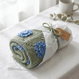 May include: A rolled-up, handmade crochet blanket in shades of green and blue, featuring floral appliques. The blanket is wrapped in white paper and tied with a green string, with a small tag that reads "For You".